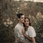 A loving couple embracing in an outdoor field, surrounded by wildflowers, during a serene sunset.